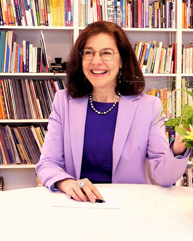 Karla Schlaepfer laughing at a table in front of a colorful bookshelf, in a lavender blazer